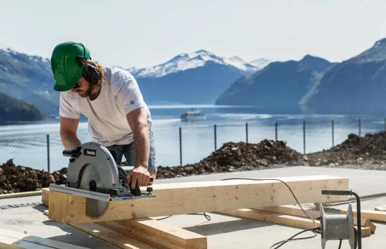 Tømrer som kapper konstruksjonsvirke med sirkelsag, med fjord og fjell bak